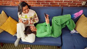 2 people sitting close on the couch, both smiling as one drinks tea and the other shows the screen of a mobile phone